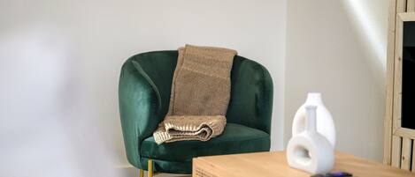 Un fauteuil en velours vert agrémenté d’un plaid tricoté, un vase blanc et une table d’appoint en bois avec des télécommandes.