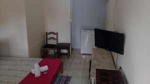 Une petite pièce comprenant un lit, un téléviseur fixé au mur, une chaise, une petite table et un réfrigérateur.