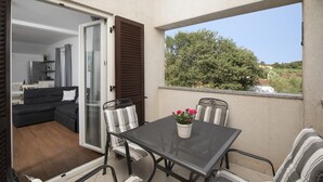 A balcony with a table and chairs, a sliding glass door leading to a living area, and a view of greenery and buildings.