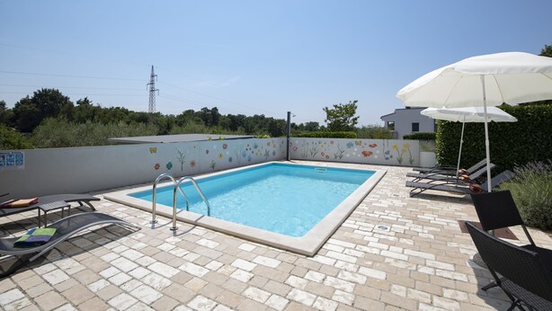 A rectangular pool with a ladder, surrounded by lounge chairs and umbrellas, on a paved patio.