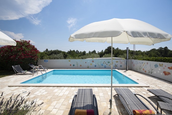 A rectangular swimming pool with a tiled deck, surrounded by lounge chairs and umbrellas, and a wall with floral murals.