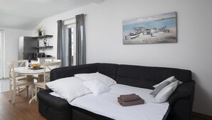 A modern living room with a black sofa, white bedding, and a painting of boats on a beach.