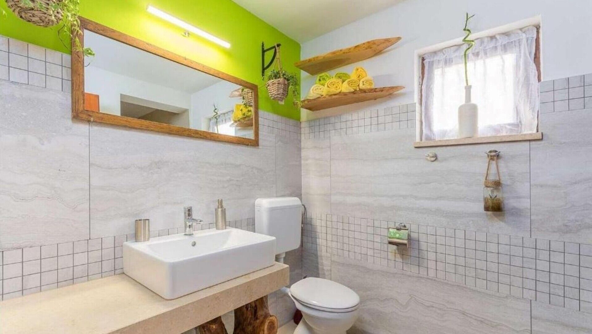 Een badkamer met een wit wastafel, toilet en spiegel. Er zijn planken met handdoeken en een raam met gordijn.