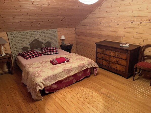 Ein gemütliches Zimmer mit Holzbalkendecke und -wänden, einem Bett mit floralem Überwurf, einem Schminktisch, einem Sessel und einem Nachttisch mit Lampe.