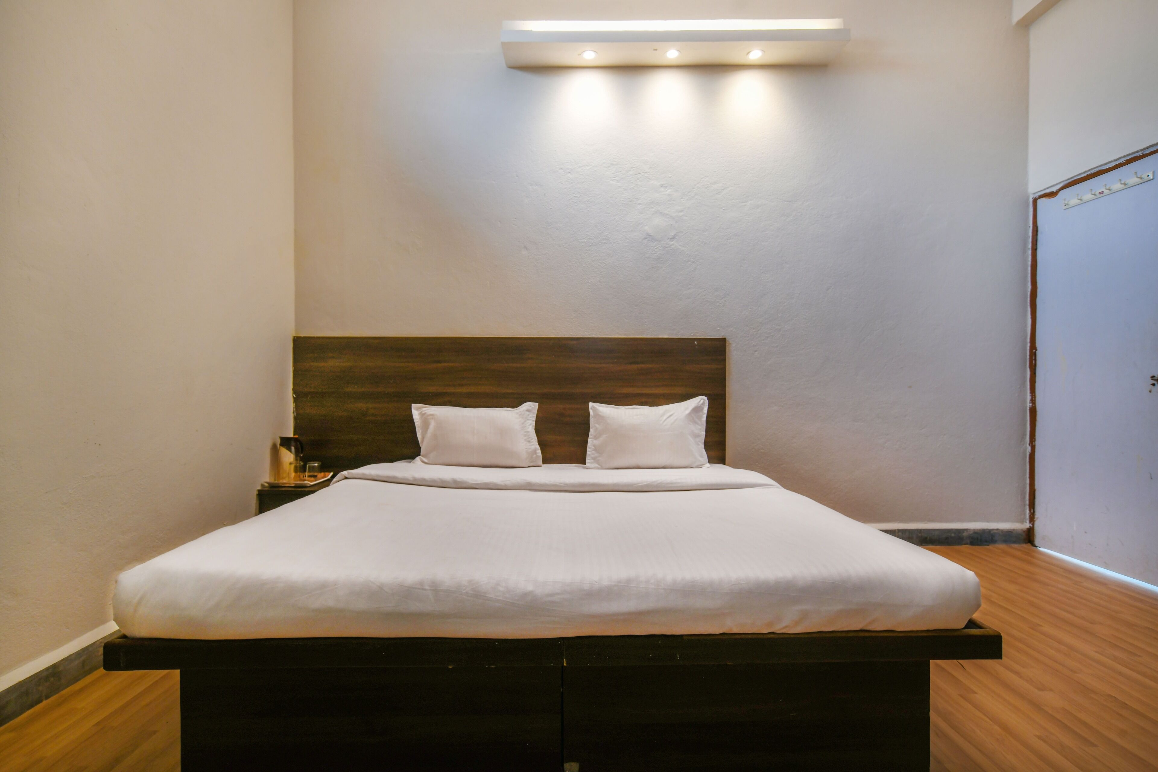 Un lit bien fait, avec du linge de lit blanc, une tête de lit en bois et une table de chevet surmontée d’une lampe.