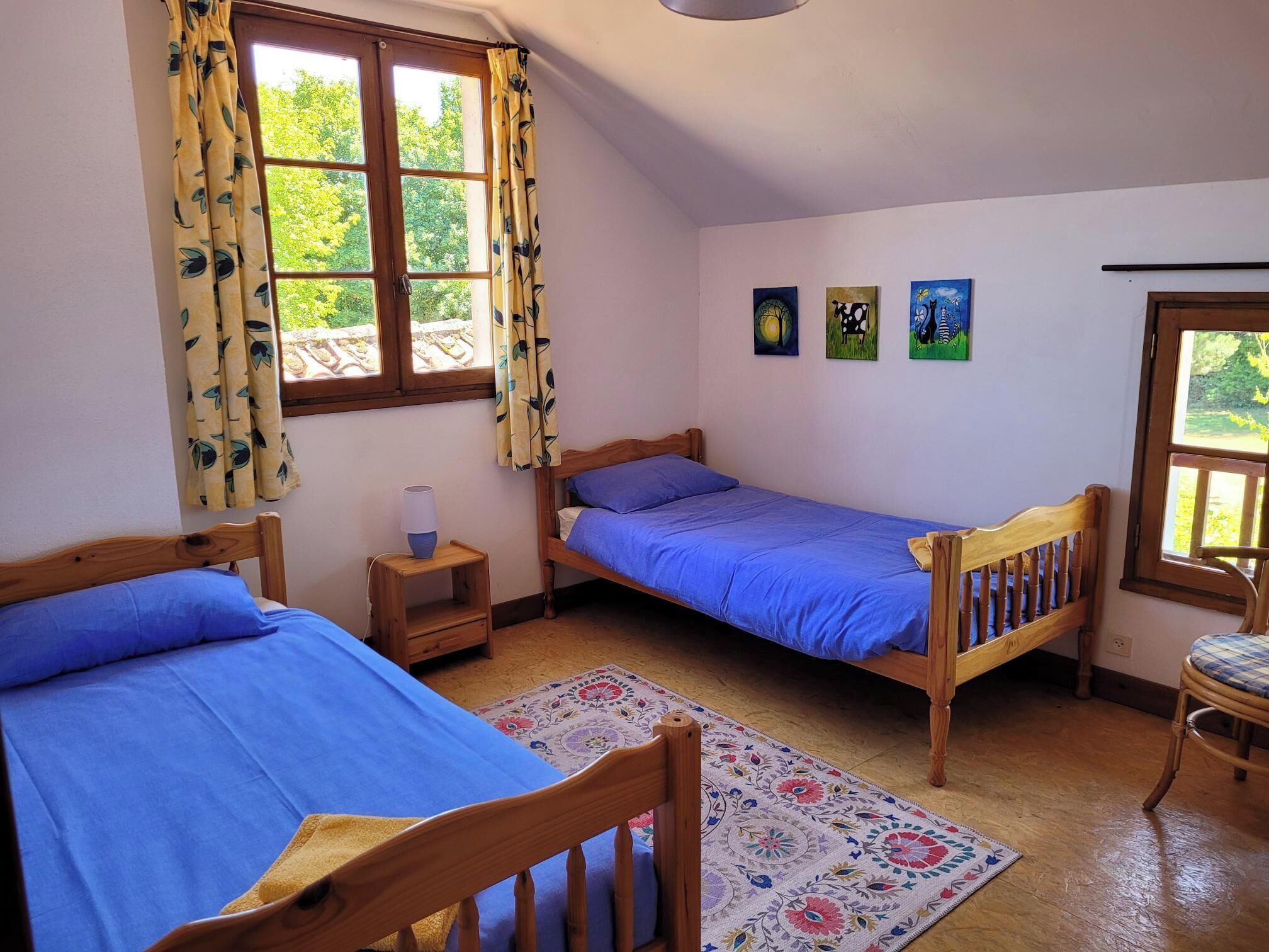 A room with two wooden beds, a small bedside table with a lamp, a chair, and a window with curtains.