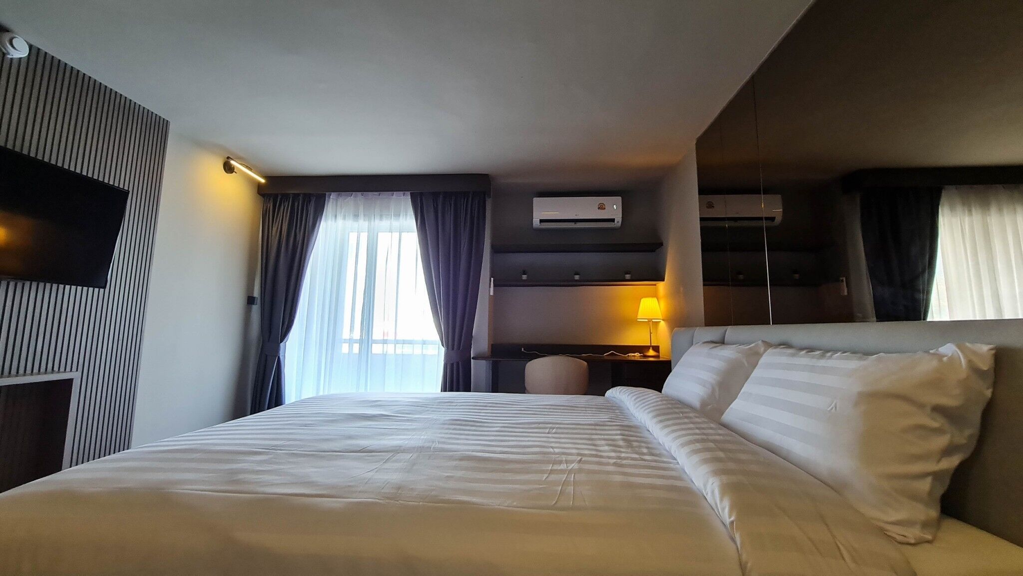 A hotel room with a large bed, a flat-screen TV, an air conditioning unit, and a window with curtains.