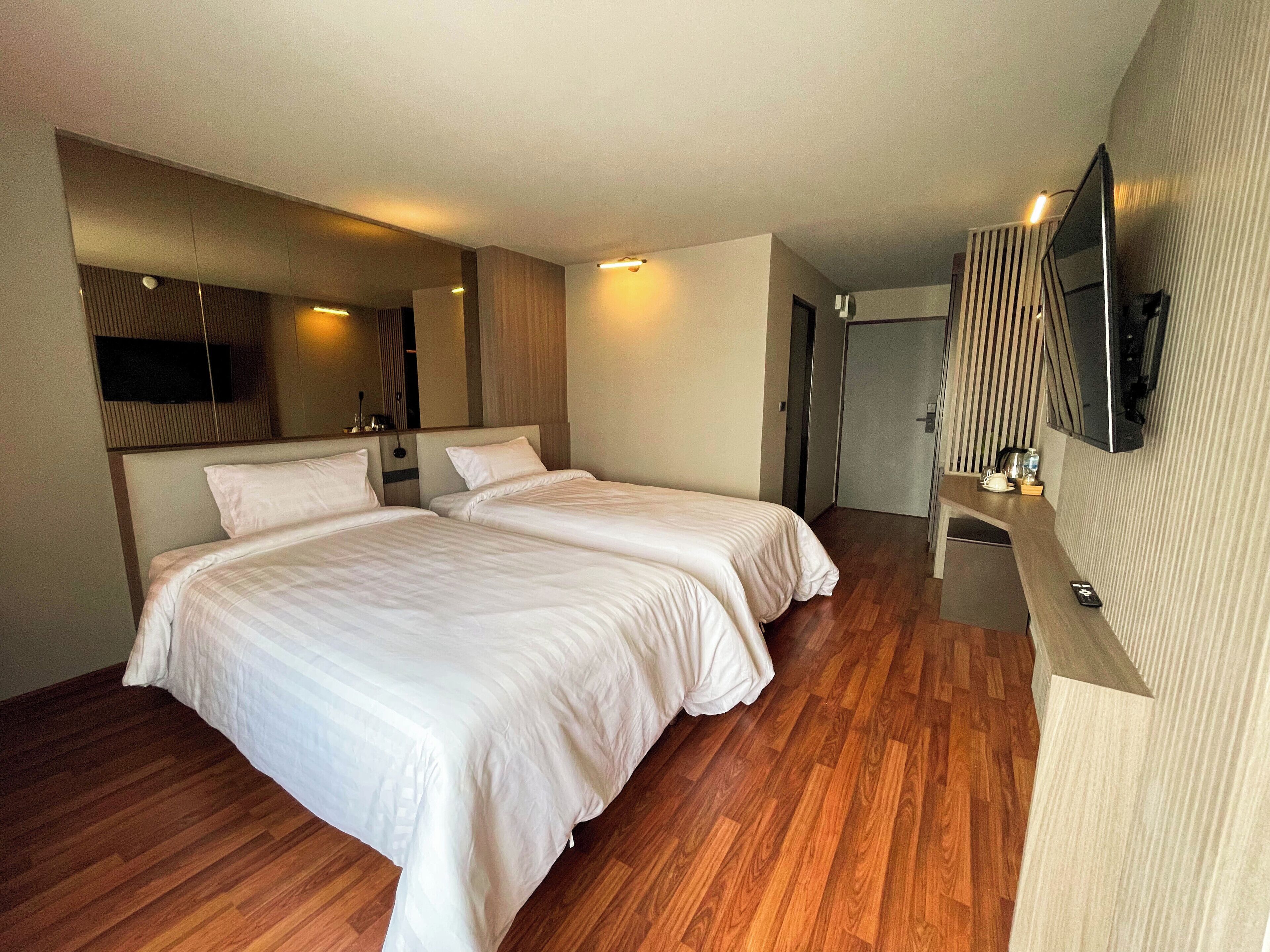 A hotel room with two beds, wooden flooring, a TV mounted on the wall, and a mirror above the bed.