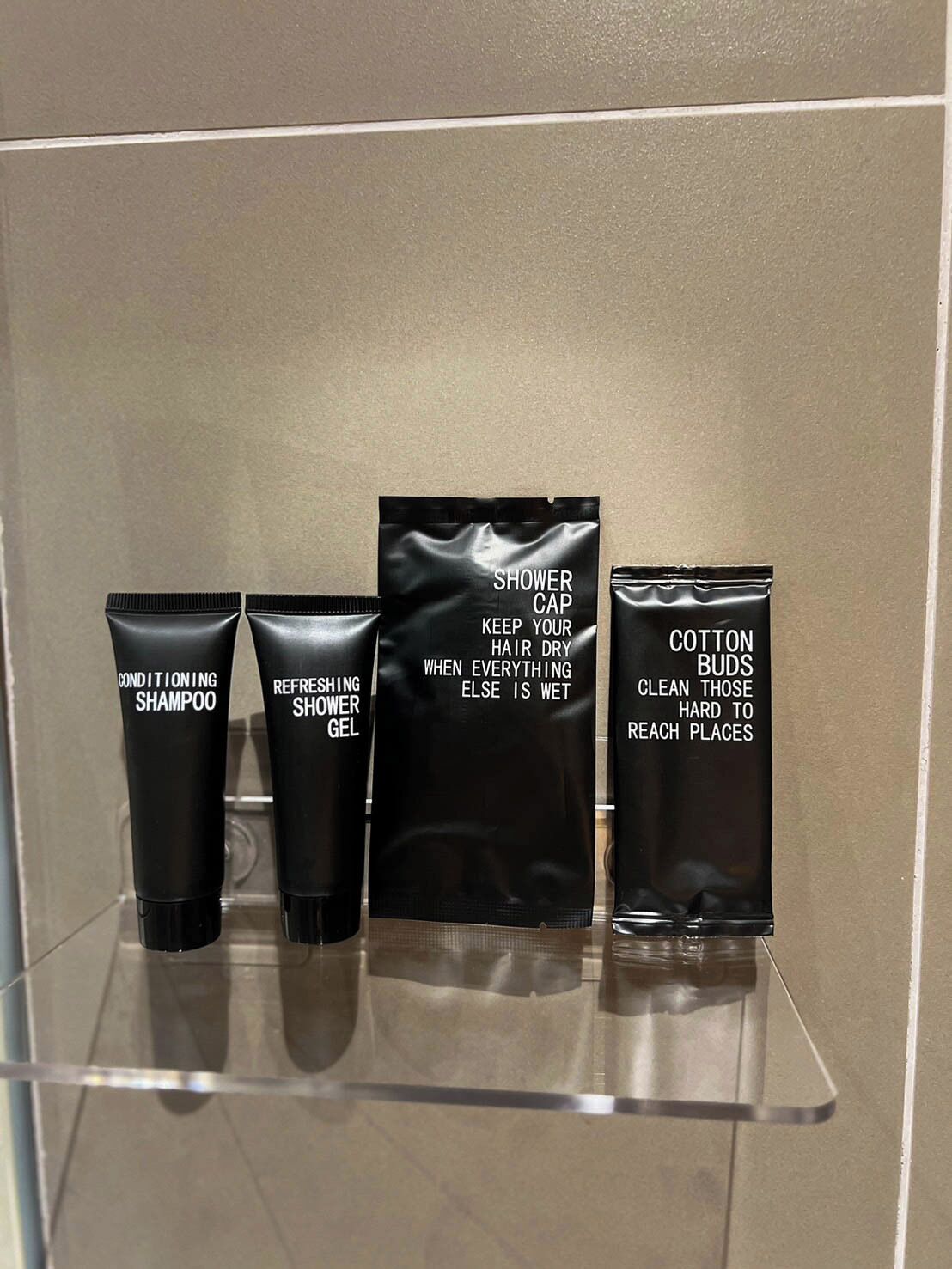 Een badkamerplank met vier zwarte toiletartikelen: een conditionerende shampoo, een verfrissende douchegel, een douchemuts en een verpakking met wattenstaafjes.