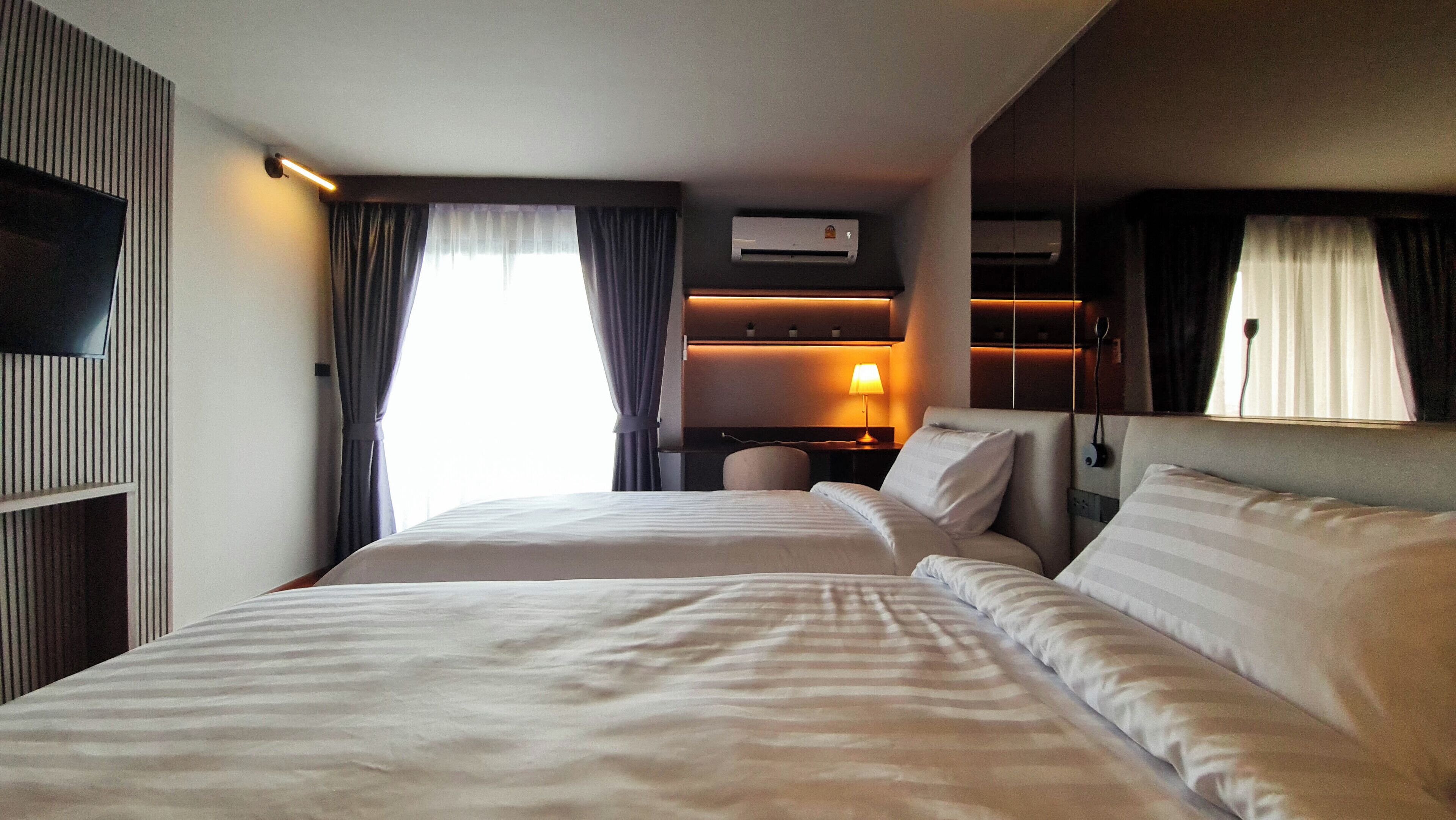 A hotel room with two beds, a TV, an air conditioning unit, a desk with a lamp, and a window with curtains.