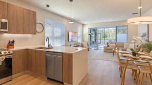 Una cocina moderna con una isla central, un comedor con mesa de vidrio y una sala de estar con sofá y televisor.