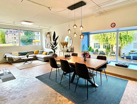 A modern living room with a dining area, large windows, and a view of the street.