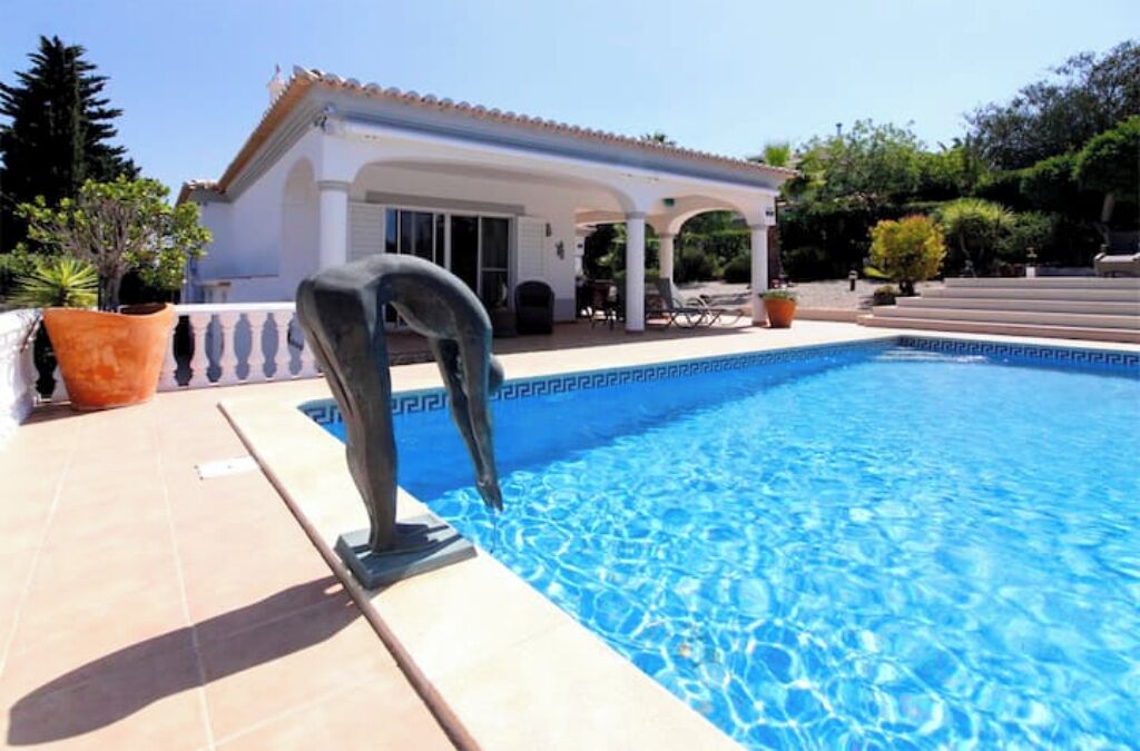 A swimming pool with a clear blue water surface, a covered patio area, and a building with a tiled roof in the background.