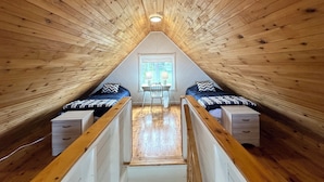 A triangular attic bedroom with wooden walls and a sloped ceiling. There are two beds with matching headboards and a small table with a chair. - Brackley Rose- Charming Water Front Cottage In Brackley, Minutes To Beaches (Brackley Beach)