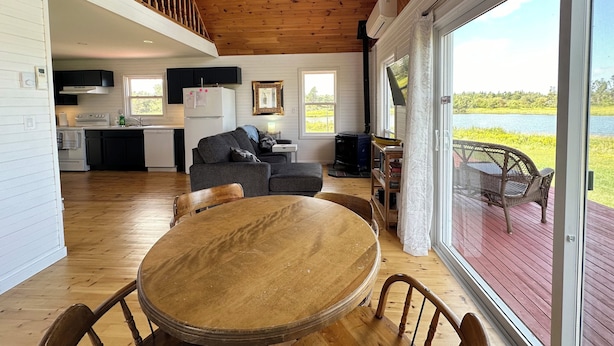 A spacious living area with a wooden ceiling, a dining table, and a view of a lake through large windows. - Brackley Rose- Charming Water Front Cottage In Brackley, Minutes To Beaches (Brackley Beach)