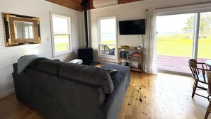A cozy living room with a sofa, a fireplace, a television, and large windows offering a view of the outdoors. - Brackley Rose- Charming Water Front Cottage In Brackley, Minutes To Beaches (Brackley Beach)
