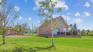 A single-story house with a red roof and a large circular window, surrounded by a well-maintained lawn and a few trees. - Brackley Rose- Charming Water Front Cottage In Brackley, Minutes To Beaches (Brackley Beach)