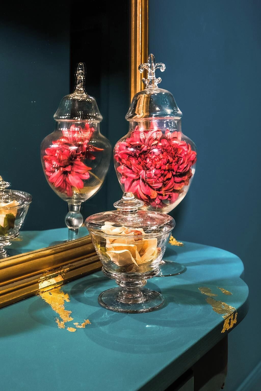 A mirror reflecting three glass vases with flowers, a small glass bowl, and a gold-framed mirror on a teal surface.