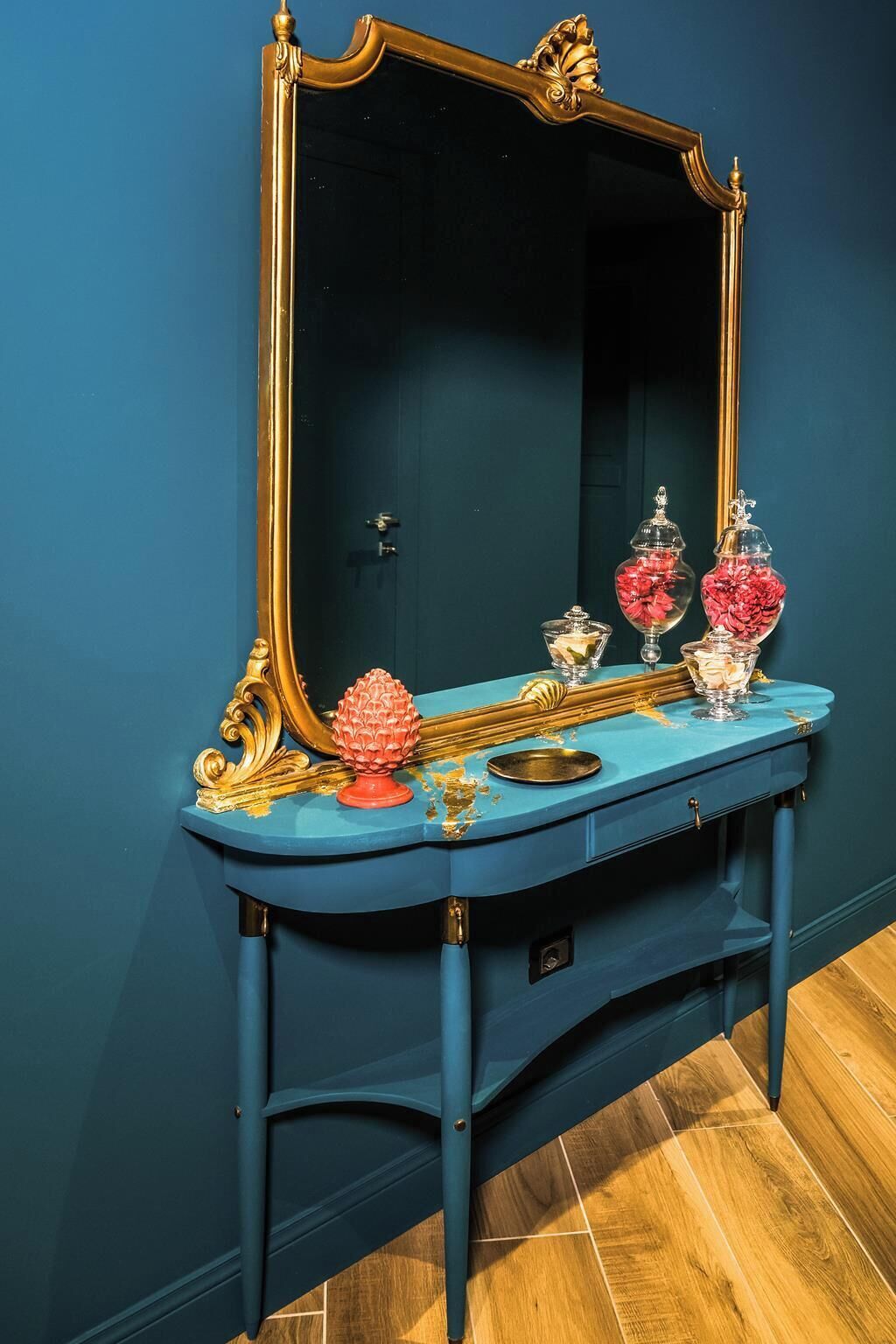 A blue vanity with a gold-framed mirror, a red decorative item, and two glass containers with pink flowers.