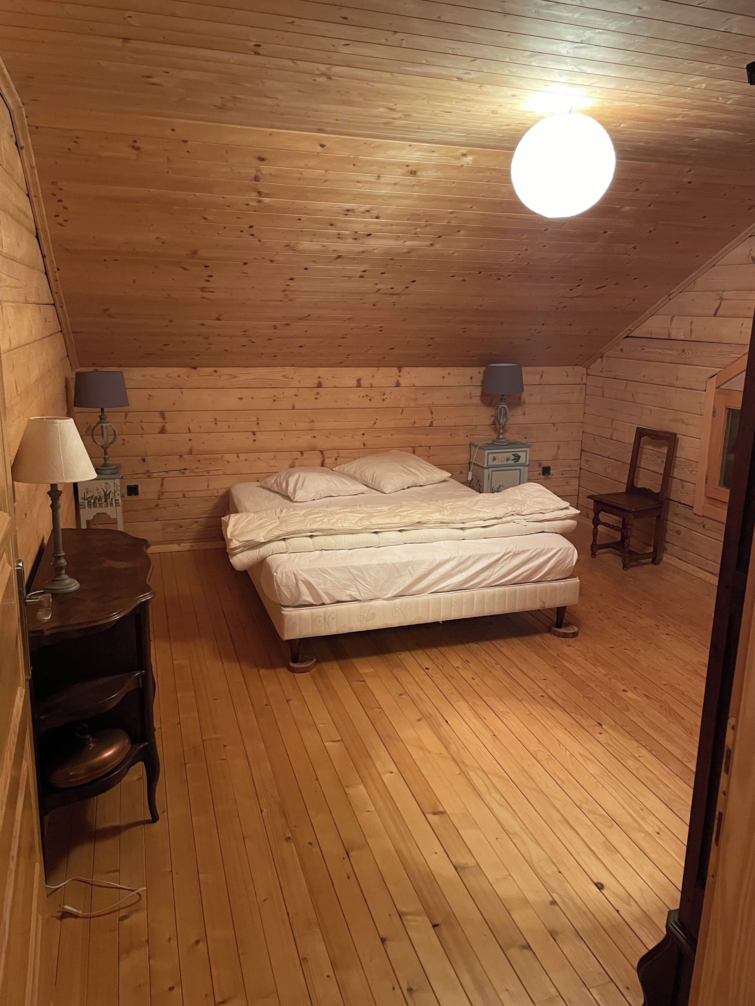 A cozy wooden cabin bedroom with a bed, two bedside lamps, a small table, and a chair.