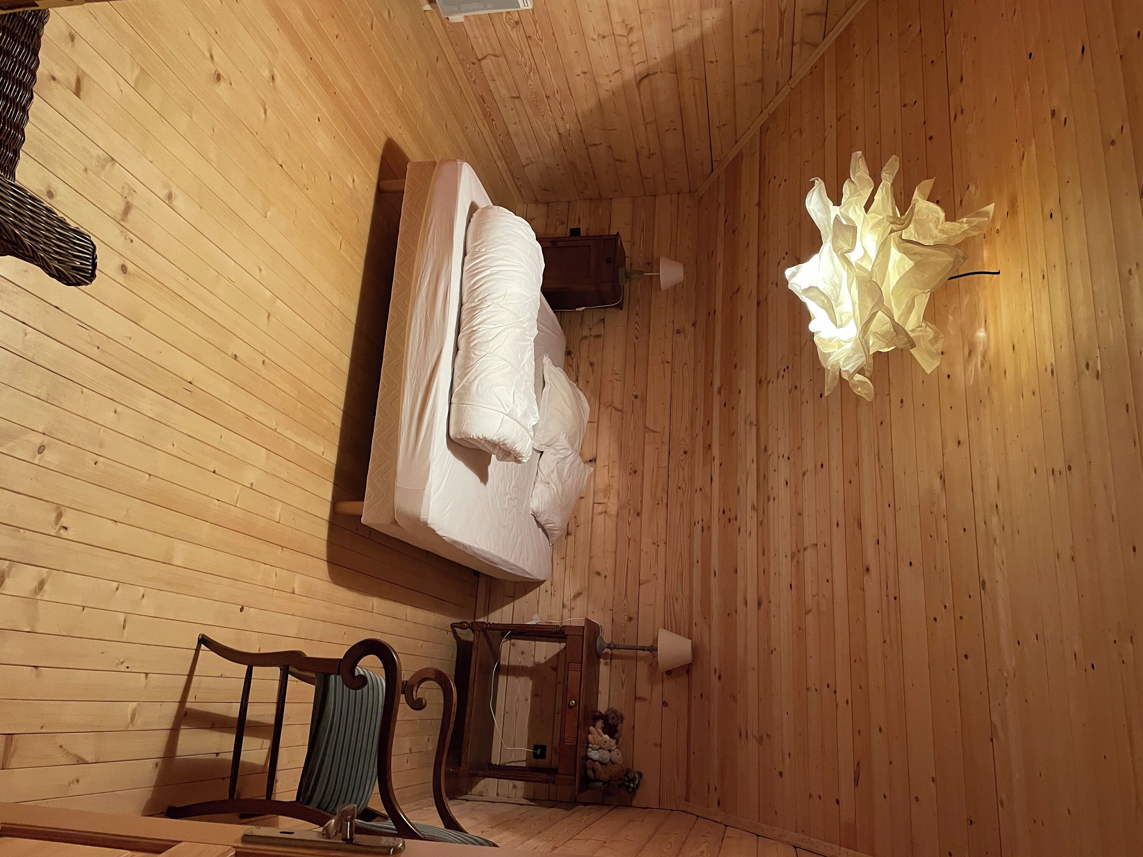 A wooden cabin room with a bed, a chair, a small table, and a lamp.