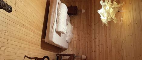 A wooden cabin room with a bed, a chair, a small table, and a lamp.