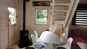 Interior acollidor d'una cabana amb una taula central, cadires blanques, un bressol de fusta negra, una finestra amb vista a la verdura i una zona al sostre accessible per una escala.