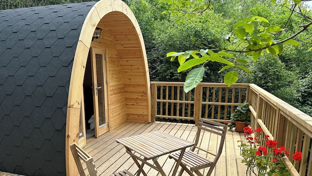 A wooden deck with a table and chairs, a potted plant with red flowers, and a wooden structure with an arched entrance. - Logement Insolite Cabane Pod sur Pilotis (Valencisse)