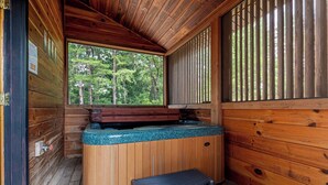 A wooden cabin with a hot tub, a window with a view of trees, and a wooden bench.