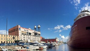 Ein großes Schiff liegt in einer Marina vor Anker, im Hintergrund sind Gebäude und eine Kuppel zu sehen.