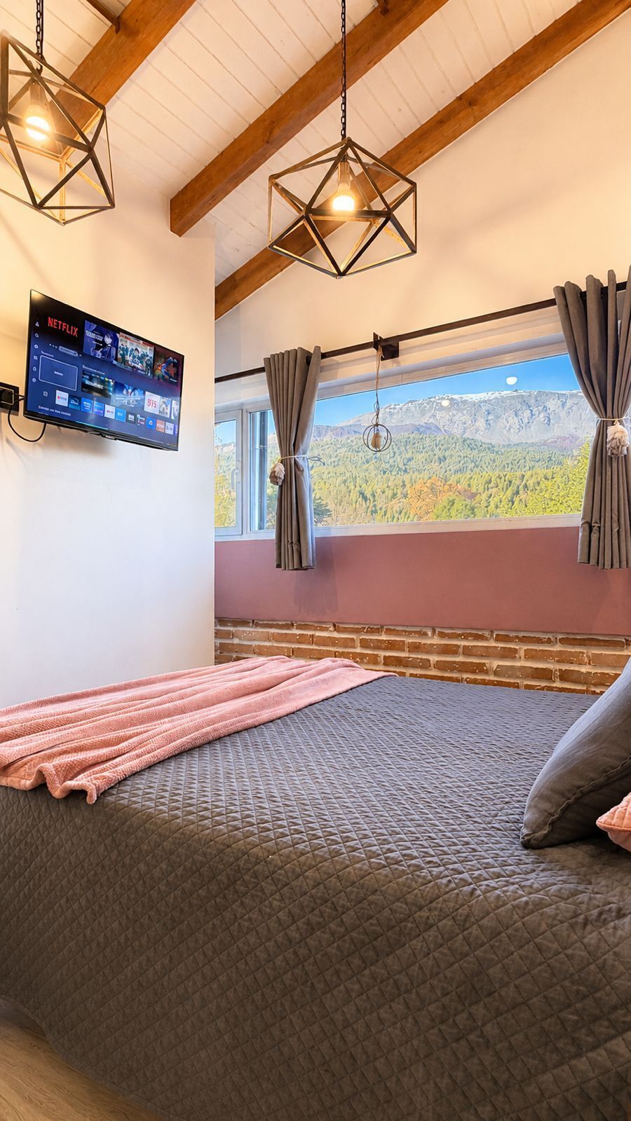 Een slaapkamer met een bed, een aan de muur bevestigde televisie, een raam met gordijnen en uitzicht op een berglandschap.