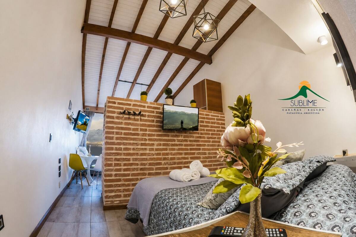 Une chambre d’hôtel avec un mur d’accent en briques, un téléviseur fixé au-dessus, un lit avec une literie à motifs, un vase avec des fleurs et une table en bois avec une télécommande.