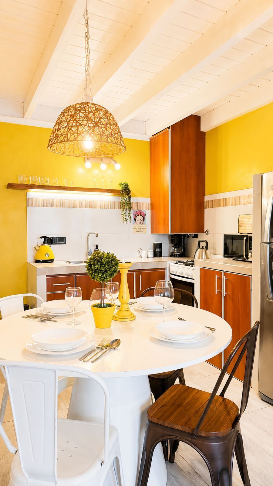 Una cocina moderna con una mesa redonda de comedor, armarios blancos y de madera, una lámpara colgante y una pared amarilla.