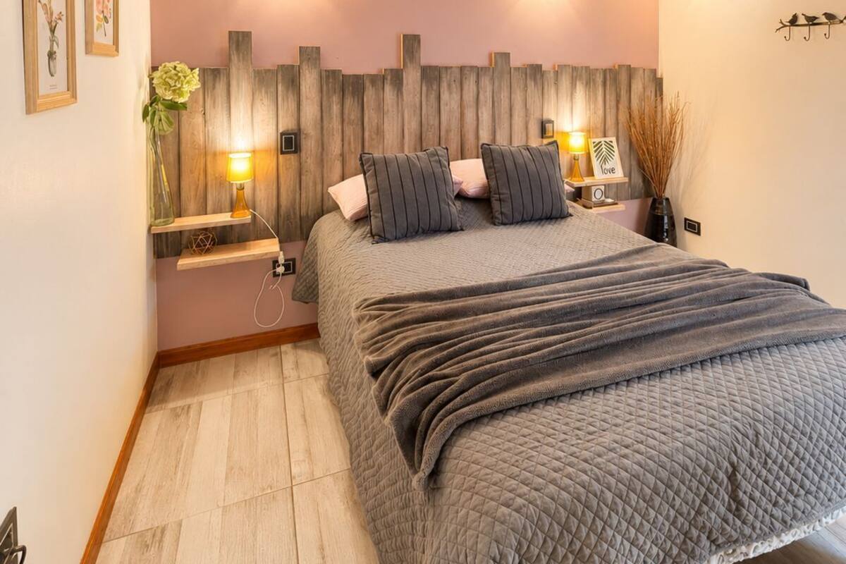 Une chambre à coucher avec un lit, des poutres en bois, des lampes fixées au mur, une table de chevet et une étagère murale ornée d’un vase.