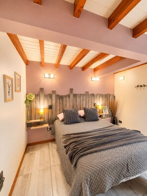 Een slaapkamer met een bed, houten balken, wandlampen, een nachtkastje en een wandplank met een vaas.