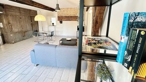 A modern living room with a glass coffee table, a grey sofa, and a dining area with a glass table and chairs. There is a bookshelf with books and a potted plant. - Gite Alcea - Château de Panisseau (Thénac)