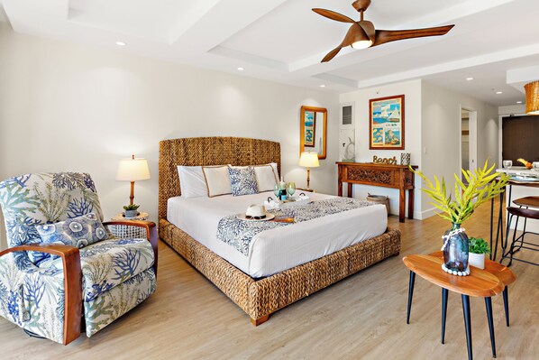 A spacious bedroom with a large bed, a wooden headboard, a wicker armchair, and a small wooden table with a vase. - Ocean View steps from beach, dining, shopping 418 (Lahaina)