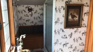 A room with a patterned wallpaper depicting horses and riders, a wooden bench, a framed picture of a horse, and a potted plant by the window.