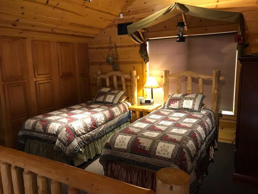 A cozy cabin-style room with two wooden beds, a small table with a lamp, and a wooden ceiling.