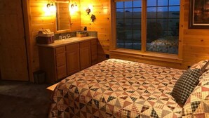 A cozy cabin bedroom with a wooden interior, a large bed with a patterned quilt, a window with a view of the outdoors, and a bathroom with a sink and mirror. - The Snow Goose Lodge (Jamestown)