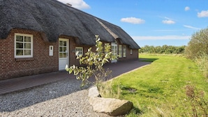 Een landelijke vakantiewoning met rieten dak, een gevel van baksteen, een grindpad en een kleine tuin met een boom en rotsen.