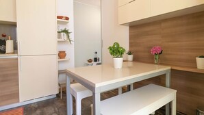 A modern kitchen with white cabinetry, a white dining table, and a bench. There are potted plants on the countertop and shelves. - Turati Family Apartment | Balcone e posto auto (Lecco)