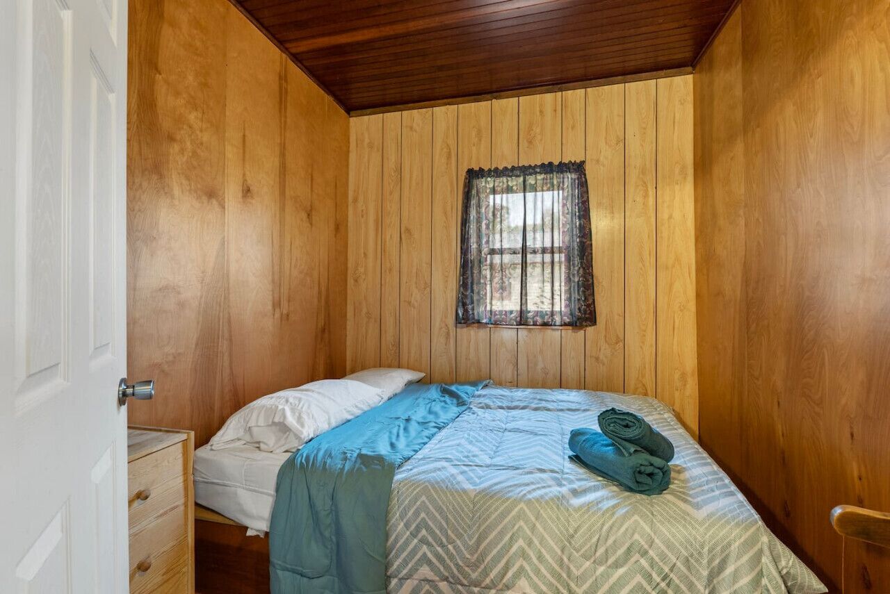 Une petite pièce aux murs lambrissés, avec un lit simple, une fenêtre ornée d’un rideau en dentelle et une commode en bois.