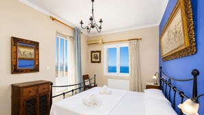 A bedroom with a bed, a wooden dresser, a chair, a nightstand, a chandelier, and a window with a view.