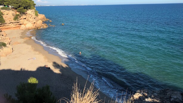 Ein Küstenstrand mit klarem, blauem Wasser, sandigem Ufer und felsigen Klippen.