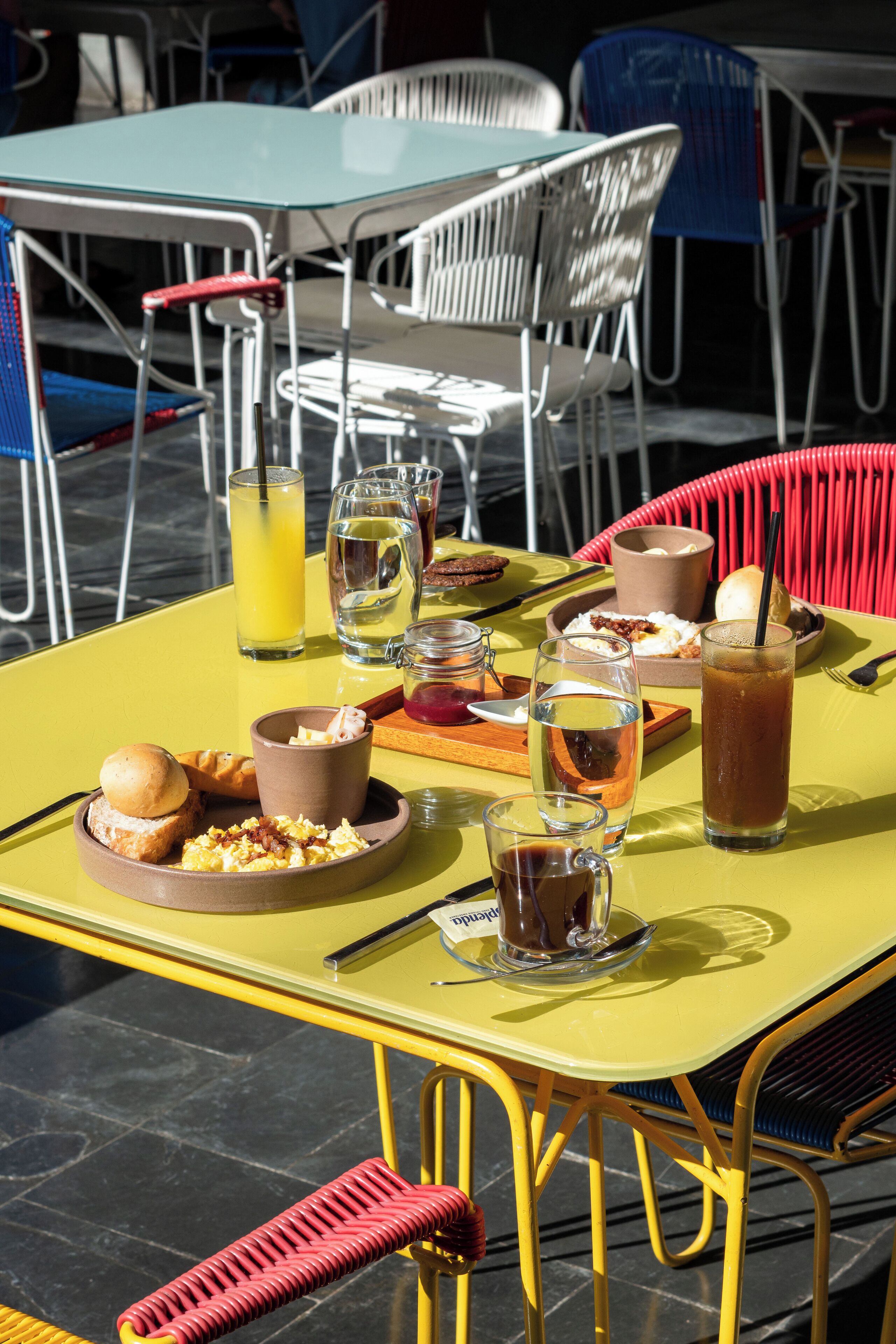A yellow table with breakfast items, including eggs, toast, and juice, set outdoors with metal chairs and a tiled floor.