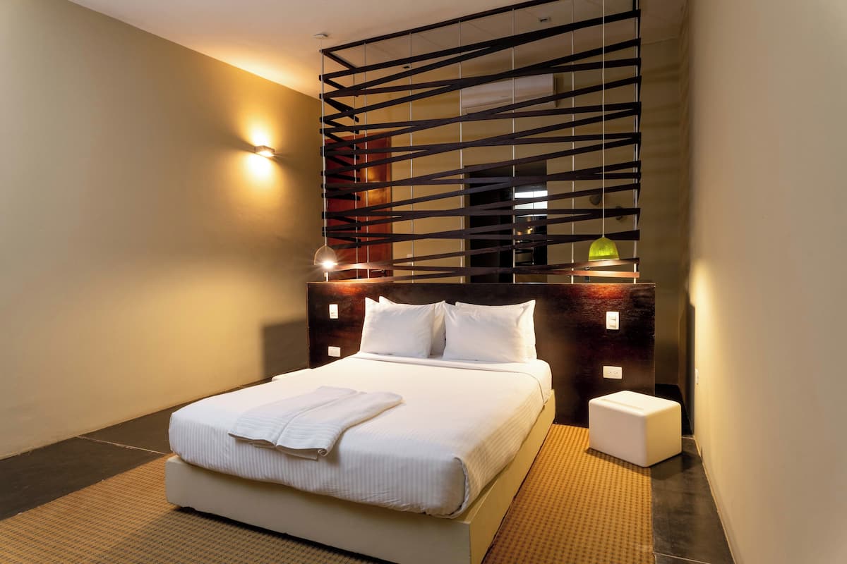 A modern hotel room with a large bed, a headboard with integrated lighting, a small bedside table, and a wall-mounted shelf.