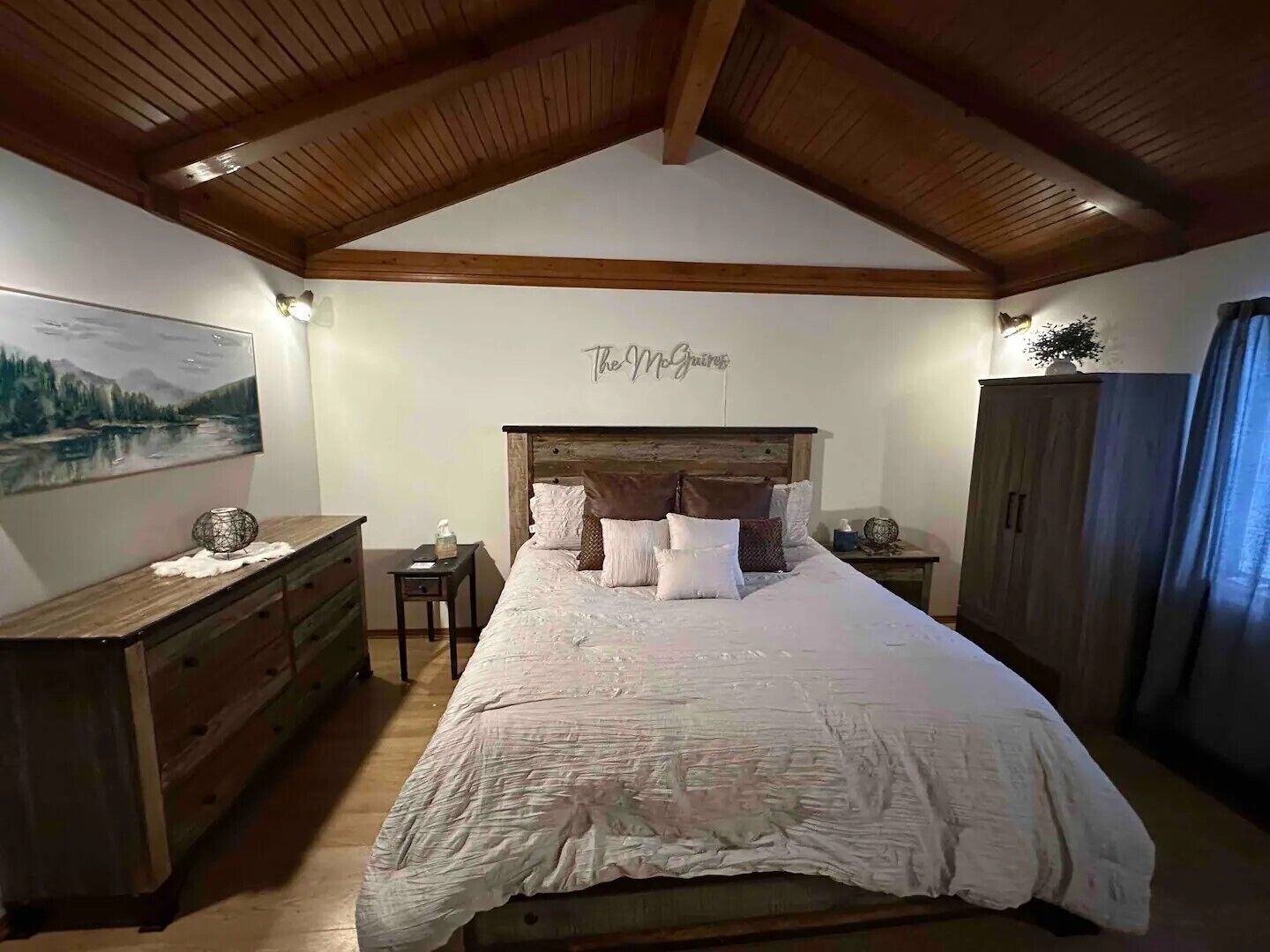 Une chambre avec un plafond en bois, un lit recouvert de plusieurs oreillers, une commode, une table de chevet et un tableau accroché au mur.
