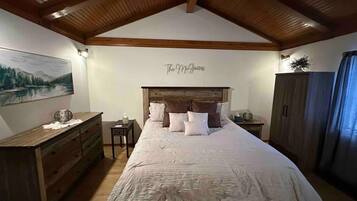 Une chambre avec un plafond en bois, un lit recouvert de plusieurs oreillers, une commode, une table de chevet et un tableau accroché au mur.