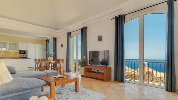 Amplia sala de estar con sofá, mesa de comedor y televisor, con vistas a un balcón frente al mar.